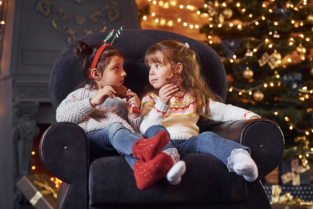 Two little girls sitting and eating cookies in christmas decorated room